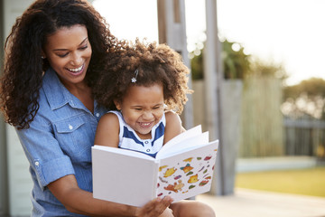 Young black girl reading book sitting on mumÕs knee outdoors