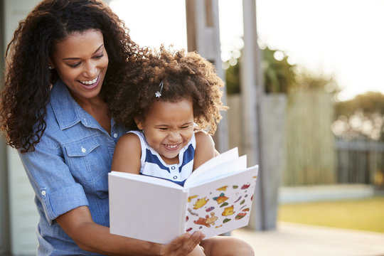 Young Black Girl Reading Book Sitting On MumÕs Knee Outdoors