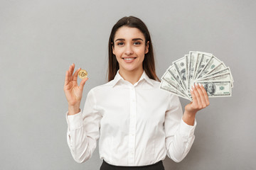 Portrait of smiling successful woman in white shirt and black skirt holding bitcoin and lots of money dollar bills, isolated over gray background