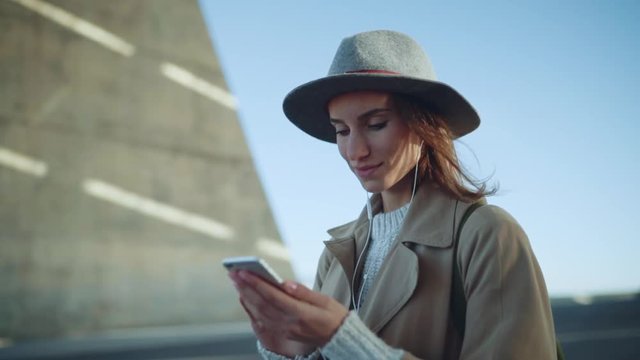 Beautiful smiling woman in hat using modern smartphone outdoor, happy smiling traveler woman wearing stylish coat enjoying music in earphones while searching sightseeing of Barcelona city, slow motion