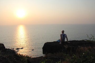 A bald man is sitting on a precipice with his back and watching the sunset or dawn against the...