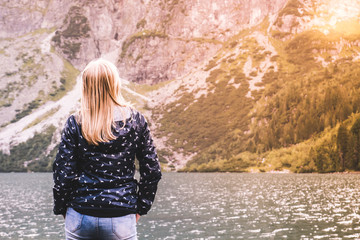 A young blonde girl stands back and looks at blue mountain lake and the mountains at sunset