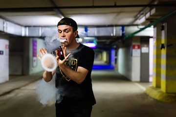 Man in cap smoke an electronic cigarette and releases clouds of vapor performing various kind of vaping tricks