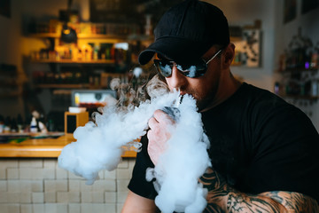 Brutal bearded man in sunglasses smoke an electronic cigarette and releases clouds of vapor at the vape shop.