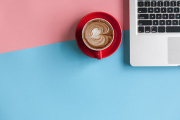 Top view of a cup of coffee and laptop with copy space on pink and blue pastel color background.