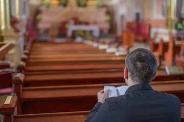 a young man or priest in a black shirt sits on a wooden bench and prays inside the Catholic Church