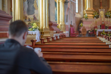 a young man or priest in a black shirt sits on a wooden bench and prays inside the Catholic Church - obrazy, fototapety, plakaty