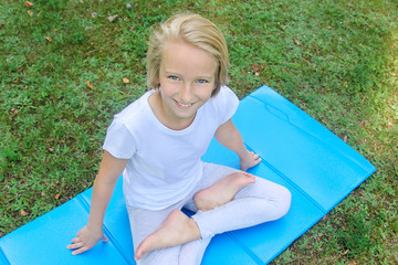 Beautiful blonde preteen girl in light clothing practicing yoga on a mat in the park. Healthy lifestyle. Outdoors workout.