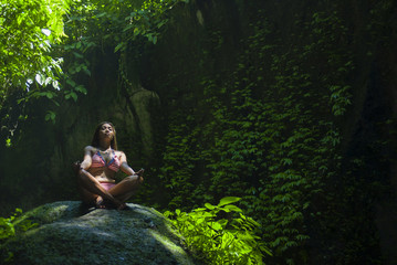 young beautiful Asian woman practicing Yoga posing sitting in lotus position meditating over a stone in a stunning natural landscape full of vegetation