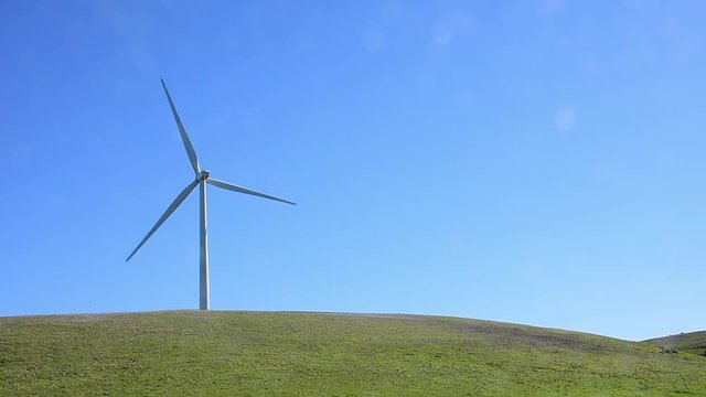 Windmill in a green hilly pasture. Windmill left one third. Camera locked.