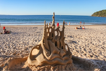 Fancy sand castle with moat in foreground on beach with people enjoying the sand and sea behind 