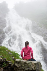 Fototapeta premium Tourist girl relaxing on rock and enjoying beautiful view of waterfall Balea in Fagarash mountains. The place of Carpathians Romania, near the Transfagarasan road. Back view