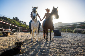 Naklejka premium beautiful caucasian couple on two horses with the sun in backlight and a dog looking them from the ground. touching hands and beautiful animals for love therapy