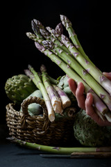 Woman hand with asparagus on textured background with artichokes in a basket 2