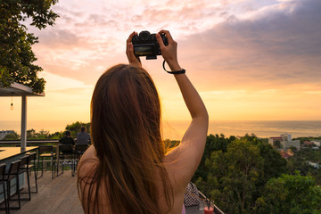 Fototapeta premium Girl with camera takes a beautiful sunset pictures over the sea from high. Back view. Average close-up