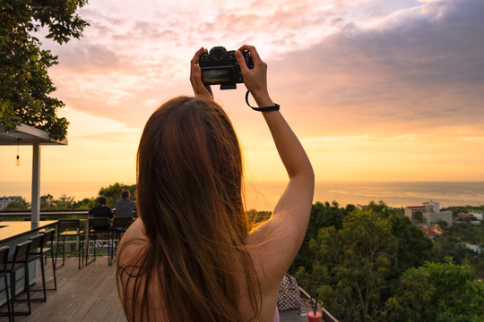 Girl with camera takes a beautiful sunset pictures over the sea from high. Back view. Average close-up