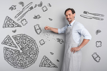 Tasty pizza. Emotional cheerful young cook standing in the kitchen and smiling while cooking tasty...