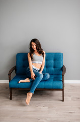 Obraz premium beautiful young brunette girl sitting on blue couch in blue jeans on gray background. Sports girl is resting