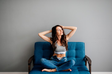 beautiful young brunette girl sitting on blue couch in blue jeans on gray background. Sports girl is resting