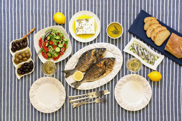 Freshly cooked seafood grilled sea bream fishes, sardines in olive oil and vegetable salad, olives, feta cheese, bread, white wine served for two persons on striped tablecloth.