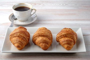 Three croissants on a rectangular plate and a cup of black coffee on a light table texture. Place for your text.