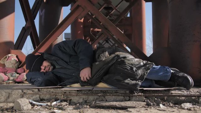 Homeless senior man in jacket sleeping outside in cold weather in city under the bridge. Senior beggar male covers his eyes with hat and tries to get warm, covering himself with jacket. Dolly shot