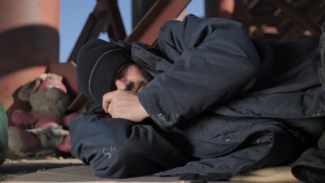 Close-up portrait of homeless beggar senior male sleeping under the bridge in jacket and hat. Cold homeless man covering eyes with hat from rising sun. Homelessness and social issues concept. Dolly