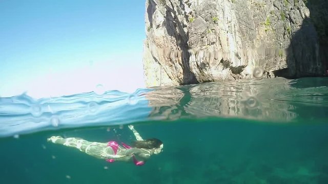 SLOW MOTION HALF UNDERWATER: Caucasian woman swims out of the deep ocean and up to the surface of cool emerald colored water. Active girl diving in the tropical sea surrounded with breathtaking cliffs