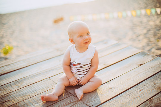 Child boy one year blond sits on a wooden dock, a pier in striped clothes, a compound near the pond on a sandy beach against a background of a river in the summer at sunset of the day