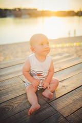 Child boy one year blond sits on a wooden dock, a pier in striped clothes, a compound near the pond on a sandy beach against a background of a river in the summer at sunset of the day