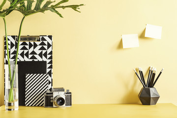 Pastel yellow desk with leafs, plant, office accessories, sticky notes and retro photo camera. Creative workspace.