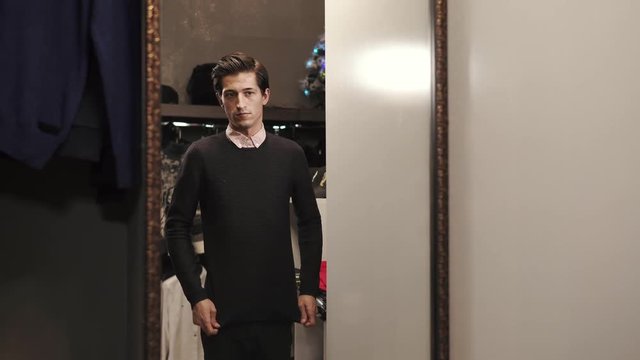 Shopper is in a fitting room of male clothes store. He is looking on his reflection in a large mirror and correcting fabric of pullover