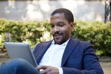 Smiling american african businessman in informal clothes working at sunny street on electronic touch pad,checking e-mail using Internet-enabled electronic device,calling friends via social networks