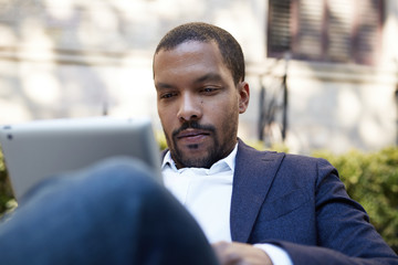 Pensive american african businessman in informal clothes working at sunny street on electronic touch pad,checking e-mail using Internet-enabled electronic device,calling friends via social networks