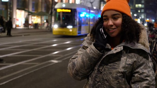 Beautiful mixed race female teenager girl young woman wearing camouflage jacket, an orange beanie hat and talking on cell phone at night with trams by Alexanderplatz Station, Berlin, Germany