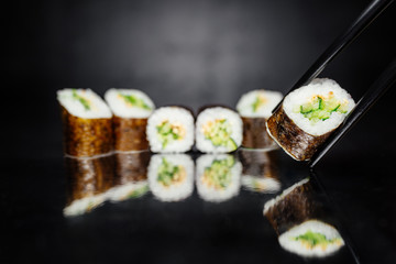Chopsticks holding roll on dark background made of Nori, Marinated rice, Sesame white, Cucumber. Traditional Japanese cuisine. Sushi menu. Japanese food. Free-space. Close-up.