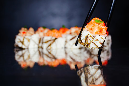 Chopsticks holding roll tobica on dark background made of nori, marinated rice, cheese, cucumber, avocado, salmon tartar, tartar from perch, spicy sauce, massago. Sushi menu. Japanese food. Free-space