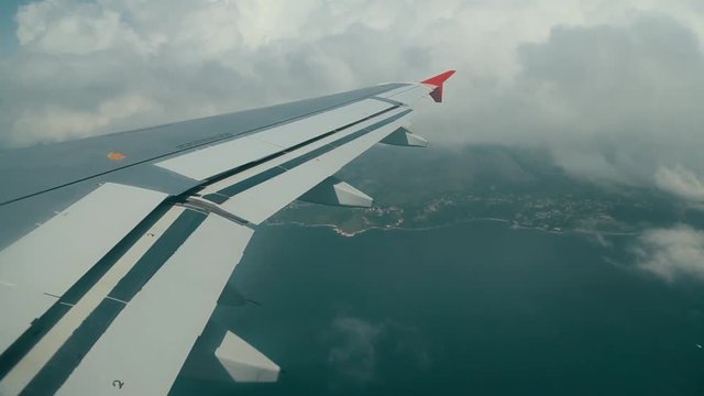View from a plane window along the wing with its red winglet as the aircraft crosses a green coastline through broken cloud. A calm and hopeful scene of a flight coming in to land at last.