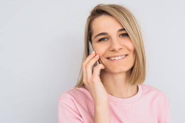 Closeup portrait of happy beautiful blonde female model, glad to speak with friend via mobile phone, posing over white wall. Pretty Caucasian woman with pleased expression talks on smartphone. People