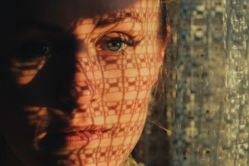 portrait of a woman standing next to a window, sunset through curtain cast a shadow on her face