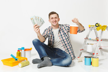 Young man holds bundle of dollars, cash money, sits on floor with paint can, instruments for renovation apartment isolated on white background. Wallpaper, gluing, painting tools. Repair home concept.