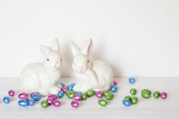 Two white ceramic rabbits with foil colored chocolate Easter eggs with copy space on white background