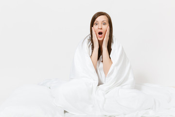 Young brunette woman sitting in bed with white sheet, pillow, wrapping in blanket on white background. Beauty female spending time in room. Rest, relax, good mood concept. Copy space for advertisement