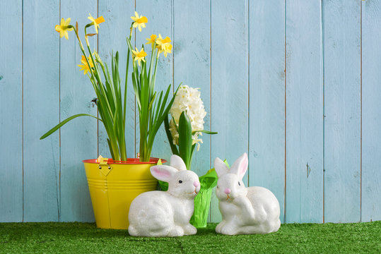 Potted daffodil and hyacinth with two ceramic rabbits on a grass surface with an aqua wood pnel background