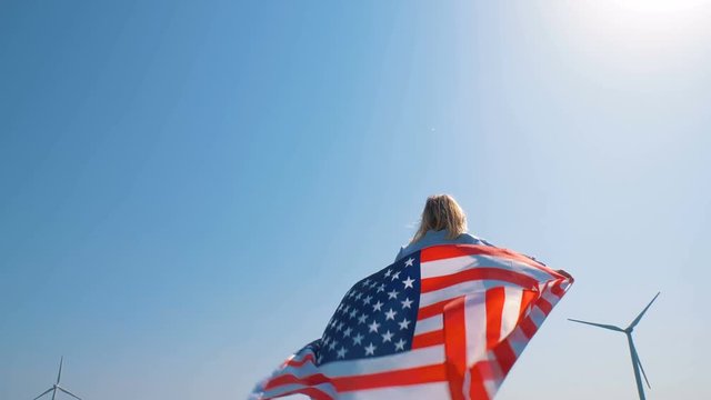 Native American girl with flag with stars and stripes near giant windmills in the field. Love to your country. Celebrating 4th July Independence Day. 4K in slow motion. American dream.