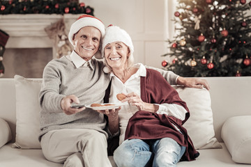 Keep smiling. Joyful mature woman feeling happiness while leaning on her husband and holding plate with biscuits