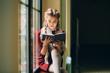 Charming woman or girl student, teenager, with cute young face and blond, two braids reading book at window on sill on dark retro background. Leisure and relaxing concept.