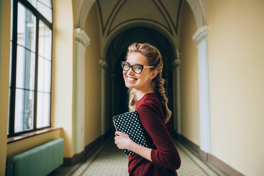 young student woman in eyeglasses carrying textbook turned around ready back to school studying. Shool girl with two braids standing indoor in hallway of old university building background.