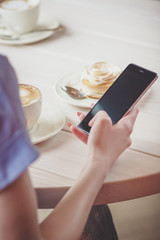 Hand with a phone, cup of cappuccino and a piece of dessert on the plate