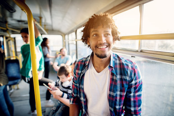 Young cheerful photogenic man is standing in the bus leaning against the window and smiling.